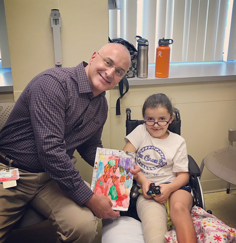 Gracie with Dr. Standard holding artwork she made for him after super knee surgery; Gracie is in a wheelchair with a cast