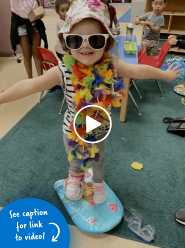 Young girl wearing big sunglasses, a beach hat, & a lei balancing on a small paddleboard inside a classroom