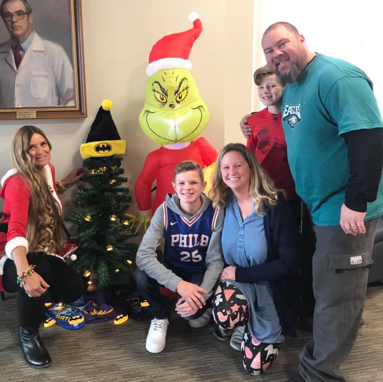 Couple with two boys and Marilyn Richardson, Pediatric Liaison, all smiling by an inflatable Dr. Seuss Grinch and small Batman-themed Christmas tree