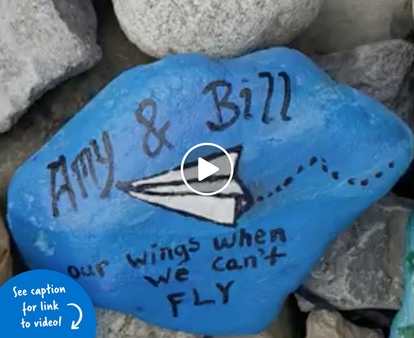 Blue rock that has a paper airplane painted on it with the words Amy & Bill - our wings when we can