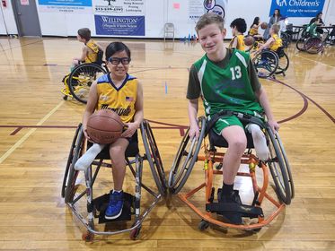 A boy and a girl who have both had rotationplasty surgery seated in wheelchairs on an indoor basketball court; the girl is holding a basketball on her lap