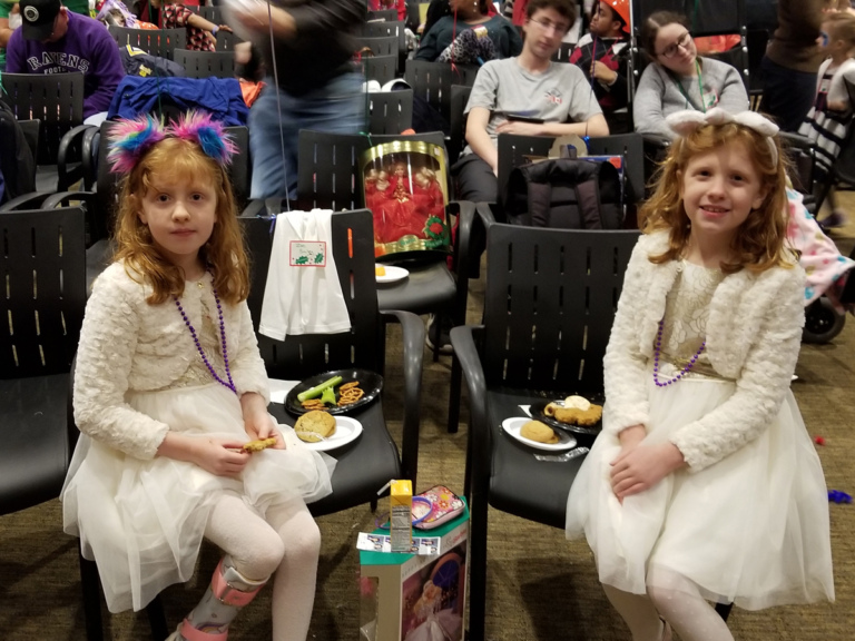 2 young girls twins, one wearing leg brace, in matching white dresses enjoying food at the International Center for Limb Lengthening pediatric holiday party