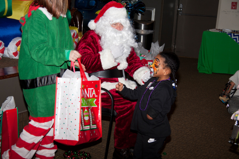 Santa and elf present a present to young girl with face painted as tiger at the International Center for Limb Lengthening pediatric holiday party