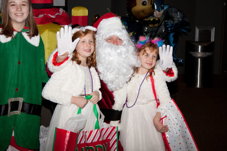 Santa and elf pose with 2 girls in white dresses with gifts at the International Center for Limb Lengthening pediatric holiday party