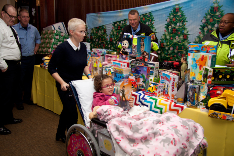 Mom wheeling young girl in wheelchair looking at toy received from firemen’s toy display table at the International Center for Limb Lengthening pediatric holiday party