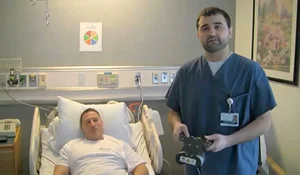 Picture from the video of research program coordinator holding the external remote control of a PRECICE internal lengthening system by the hospital bed of a male patient