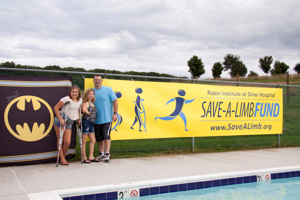 A father poses with 2 young girl patients, one using crutches, in front of Batman and Save-A-Limb Fund banners at Rubin Institute for Advanced Orthopedics 2018 Save-A-Limb Pool Party event