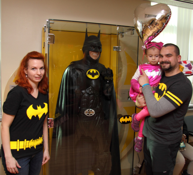 Parents in Batman shirts with father holding young girl dressed in pink Batgirl costume in front of the glass case with Lenny B. Robinson’s Batman suit at the International Center for Limb Lengthening’s Leonard B. “Batman” Robinson Memorial Valentine’s Day Party