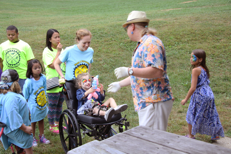 Young girl with face paint in wheelchair with external fixator on leg being handed a balloon animal at Rubin Institute for Advanced Orthopedics 2016 Save-A-Limb Fund Event