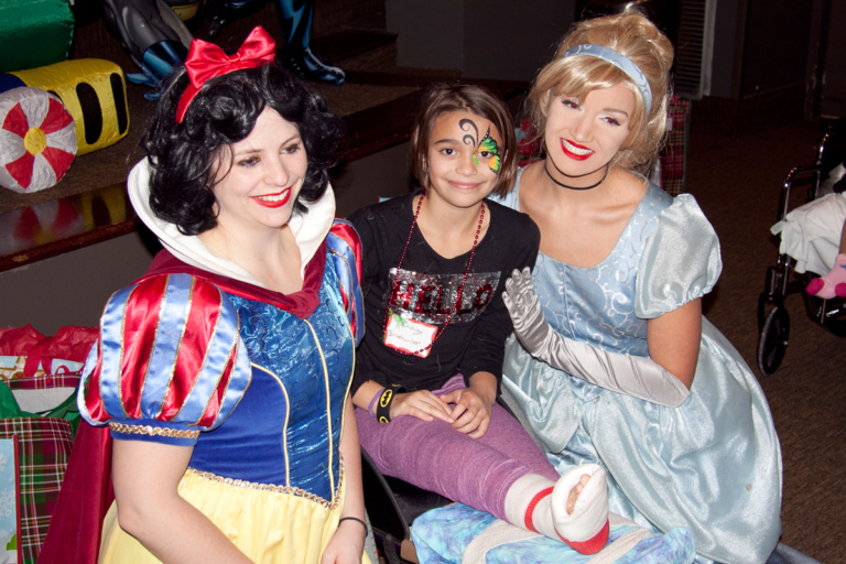 Girl patient in leg cast poses with Snow White and Cinderella at the International Center for Limb Lengthening pediatric holiday party