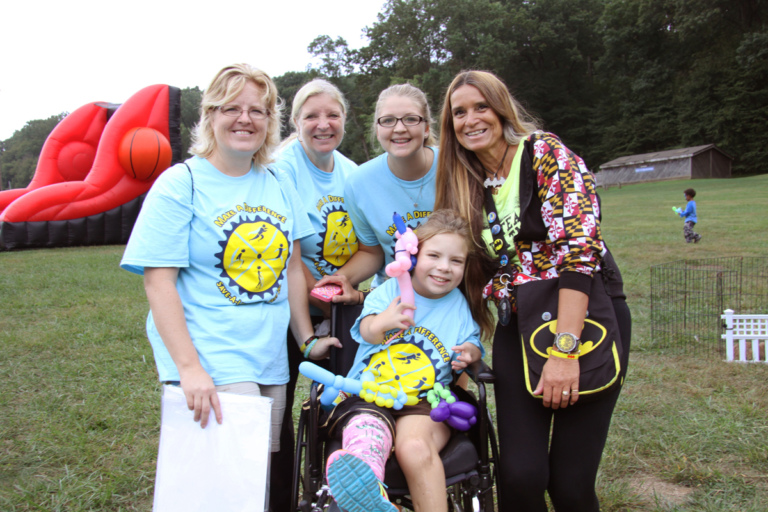 Pediatric Liaison Marilyn Richardson poses for a picture with a young girl in a cast in a wheelchair holding balloon animals and 3 participants at Rubin Institute for Advanced Orthopedics 2016 Save-A-Limb Fund Event