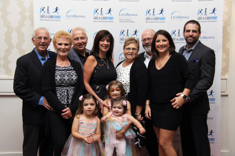 8 adult guests with 3 young girls all wearing pastel dresses standing in front of Save-A-Limb Fund photo backdrop at Rubin Institute for Advanced Orthopedics 2016 Save-A-Limb Fund Dinner