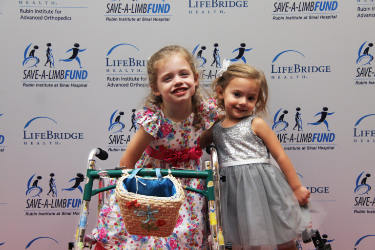 2 young girls in dresses, one with a walker standing in front of Save-A-Limb Fund photo backdrop at Rubin Institute for Advanced Orthopedics 2016 Save-A-Limb Fund Dinner