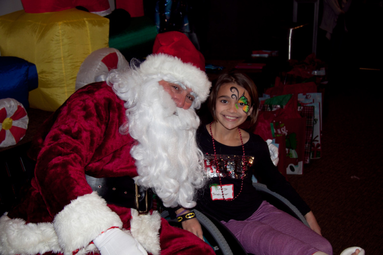 Young girl with face paint in wheelchair with cast takes picture with Santa at the International Center for Limb Lengthening pediatric holiday party