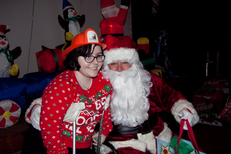 Girl on crutches and with a fireman hat on takes picture with Santa at the International Center for Limb Lengthening pediatric holiday party