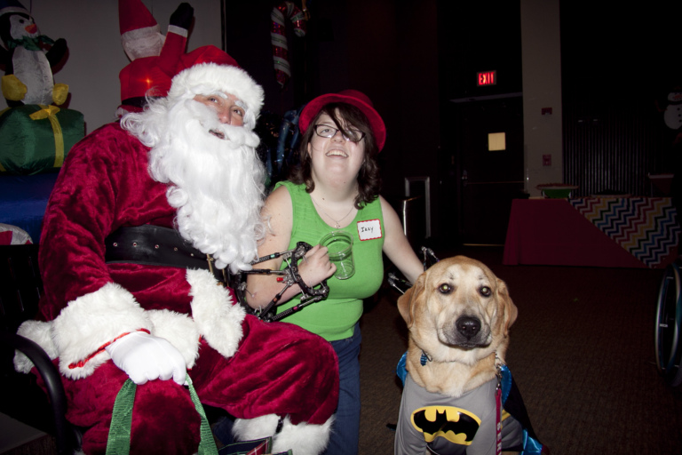 Girl with fixators on both arms takes photo with Santa and service dog dressed as Batman at the International Center for Limb Lengthening pediatric holiday party