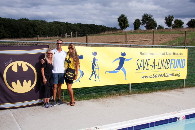 Pediatric Liaison Marilyn Richardson with two family members of Lenny B. Robinson (aka Batman) in front of Batman and Save-A-Limb Fund banners at Rubin Institute for Advanced Orthopedics 2018 Save-A-Limb Pool Party event