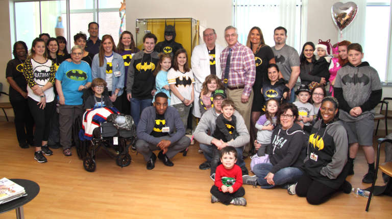 Dr. Shawn Standard and Dr. John Herzenberg pose with a large group of patients, family members and staff, mostly wearing Batman shirts, in front of the glass case with Batman costume at the International Center for Limb Lengthening’s Leonard B. “Batman” Robinson Memorial Valentine’s Day Party