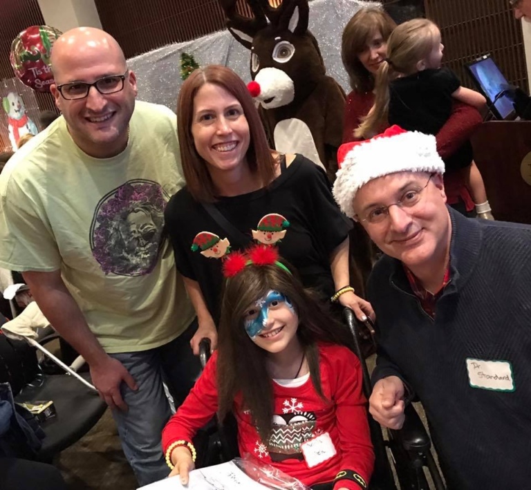 Dr. Shawn Standard in Santa hat, mother, father and young girl with face painted in a wheelchair at the International Center for Limb Lengthening pediatric holiday party