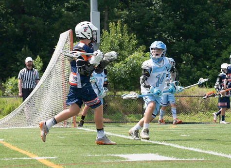 CJ in uniform number 14 with a lacrosse stick playing defense against a player on the opposing team near the goal on the playing field