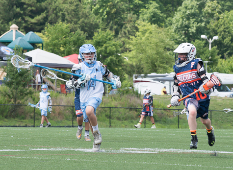 CJ in number 14 uniform running with a lacrosse stick opposite a player on the opposing team on the playing field