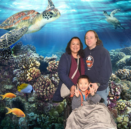 Primo smiling in wheelchair with his parents in front of a National Aquarium backdrop with coral reef, fish and turtles