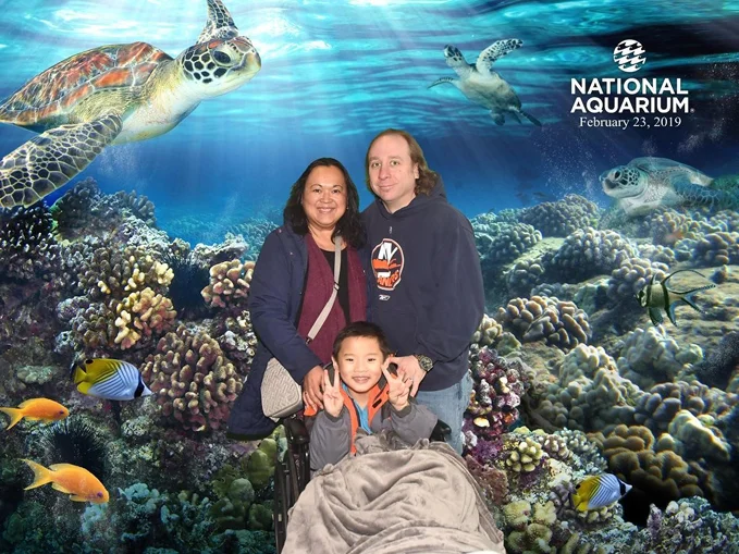 Young boy patient smiling in wheelchair with smiling parents in front of a National Aquarium backdrop with coral reef, fish and turtles