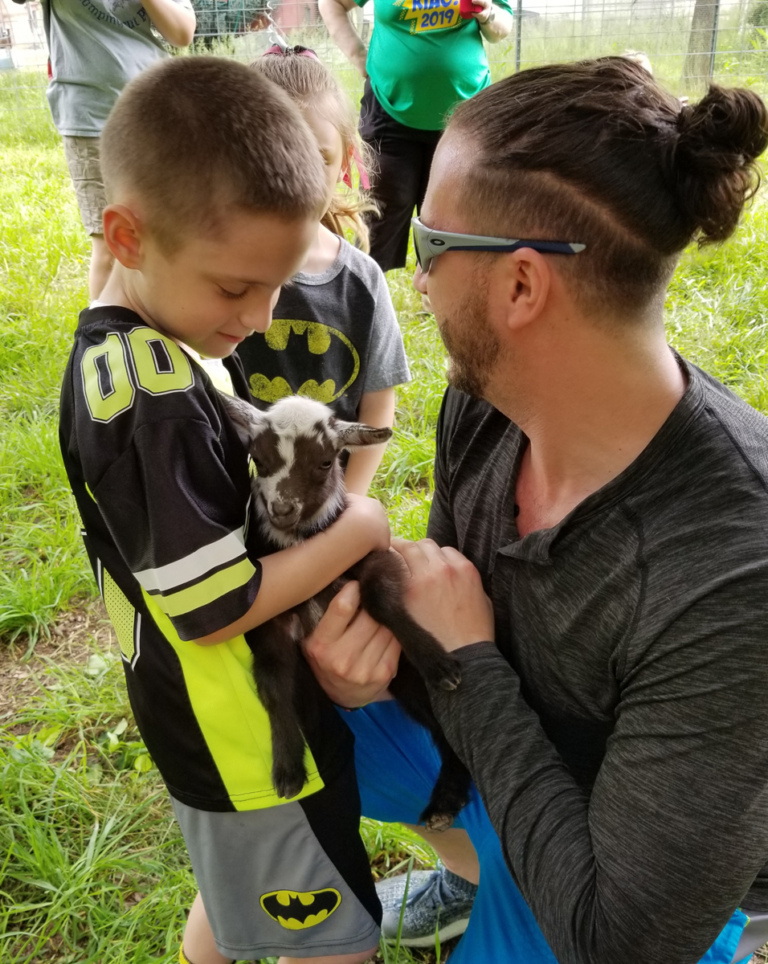 A young boy patient petting a tiny, two-week old goat that is being held by a sponsor team member