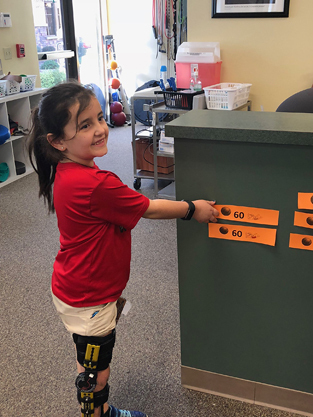 Gracie smiling while wearing her leg brace and reaching for an orange slip with the number 60 on it signifying that she hit her 60% range of motion goal at physical therapy