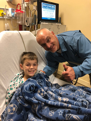 Preston smiling in a hospital bed being visited by Dr. Standard who is making a thumbs up sign