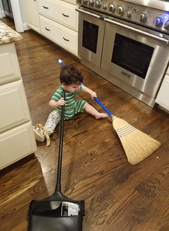 Ross wearing a cast sitting on the kitchen floor playing with a broom and dustpan