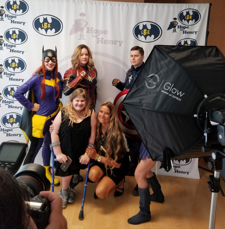 Girl patient with forearm crutches posing with 4 superheroes and Pediatric Liaison Marilyn for professional photographer in front of LBR Hope for Henry backdrop