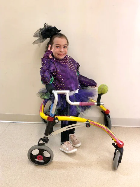 Alena in a purple and black tulle costume making a peace sign and using a wheeled-gait trainer walking aid