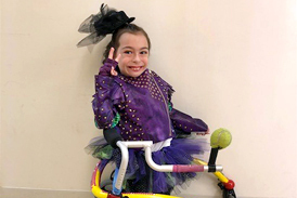 Alena in a purple and black tulle costume making a peace sign and using a wheeled-gait trainer walking aid