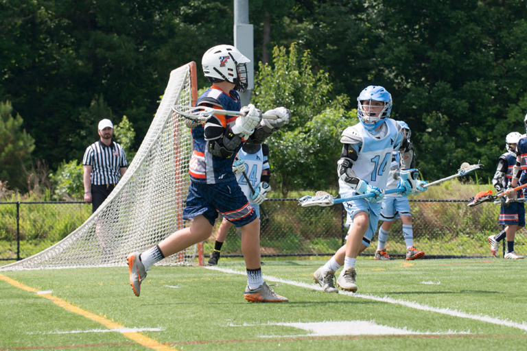 CJ in uniform number 14 with a lacrosse stick playing defense against a player om the opposing team near the goal on the playing field