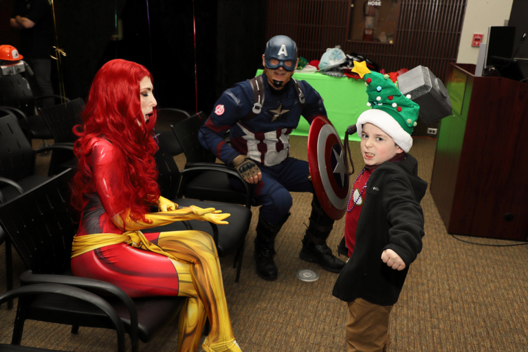 Young boy making fierce face while holding a comic villainess’ weapon next to her and smiling Captain America