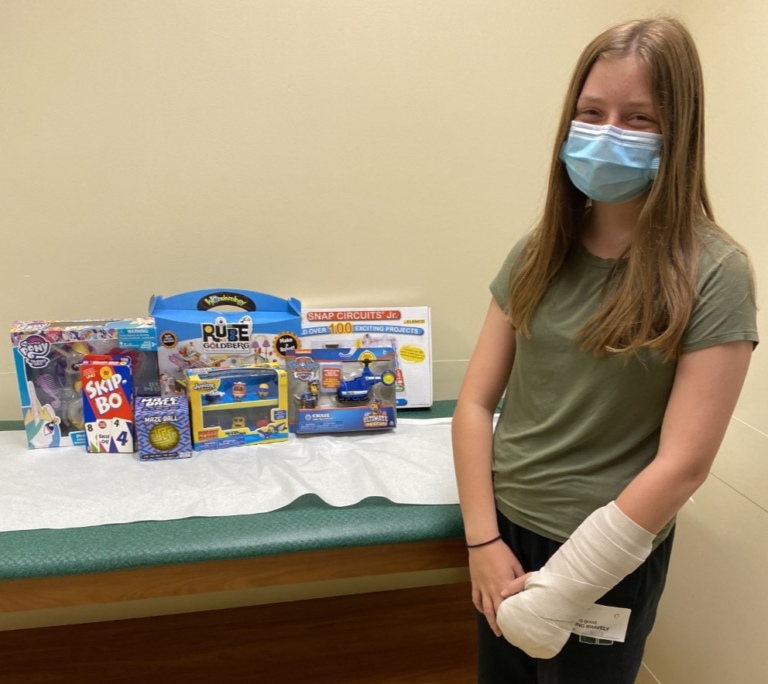 Eden with a cast on her arm standing next to toys she is donating to other patients