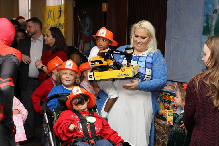 Mother holding little boy wearing orange firefighter hat and Deere Big Scoop truck with four other children wearing firefighter hats