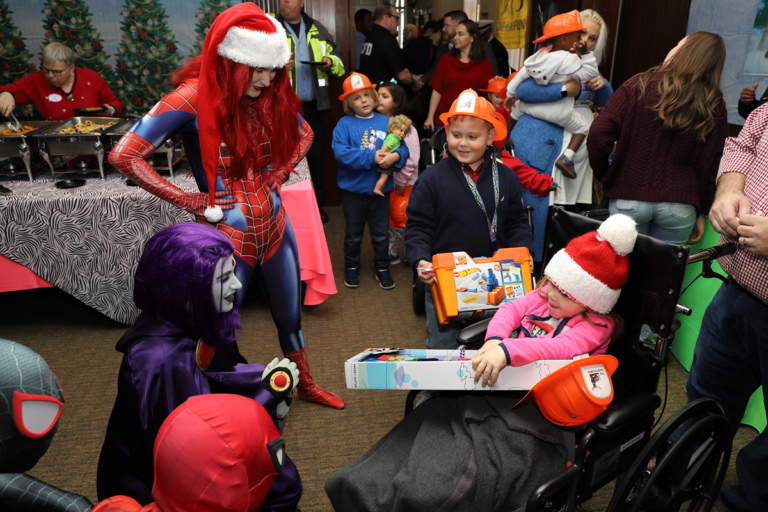 Girl in wheelchair with toy talking with comic book superheroes and villains with children with toys and firefighter hats in the background