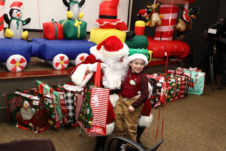 Boy in Christmas tree hat smiling on Santa’s lap getting a gift bag