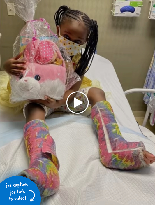 A girl with colorful casts on both her legs holding an Easter basket while laying in a hospital bed