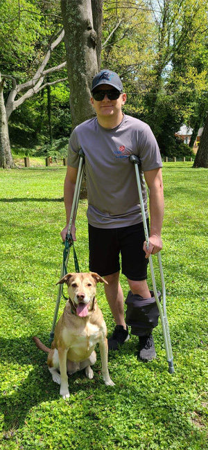 Andrew wearing a covered external fixator and using crutches standing with his dog at a park 2 weeks after surgery