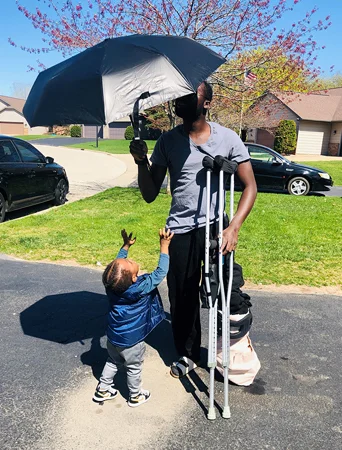 Ebou on crutches while holding an umbrella and walking with his young son