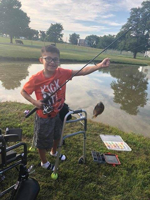 JJ catching a fish while using a walker with his wheelchair in the background