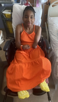Kennedee sitting in a wheelchair by a hospital bed wearing an orange dress after surgery