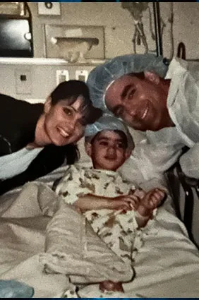 Ken as a very young boy on a hospital bed with his parents on either side before going into surgery